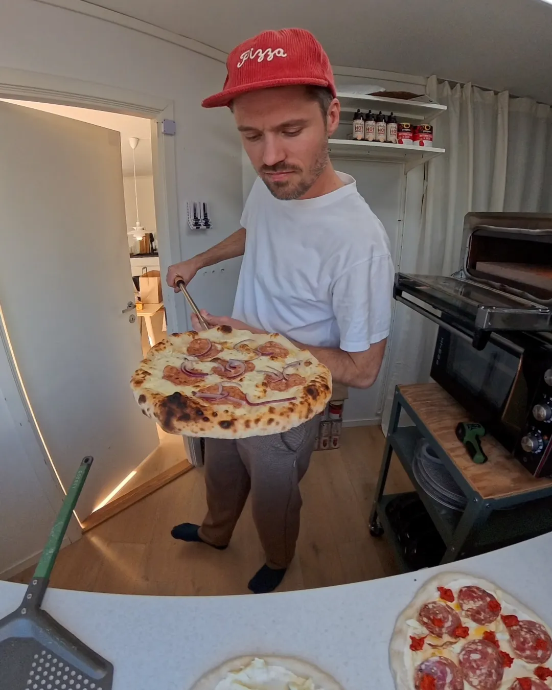 Vegard i rød pizzacaps holder en nystekt pizza på spaden, med flere pizzaer klare på benken