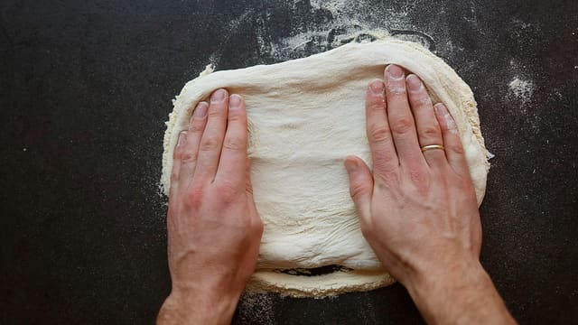 Hender som presser ut pizzadeig på en melet benk